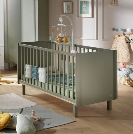 A khaki-colored cot bed in a nursery room, with height-adjustable slat base, railings, and a mobile hanging above it. The bed is accompanied by a bed mattress, not included in the purchase.