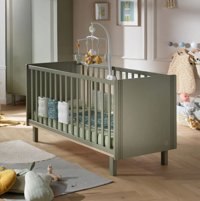 A khaki-colored cot bed in a nursery room, with height-adjustable slat base, railings, and a mobile hanging above it. The bed is accompanied by a bed mattress, not included in the purchase.