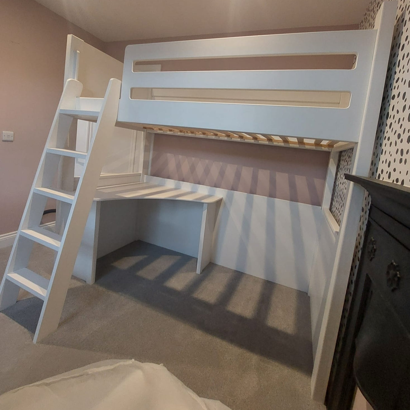 A white wooden loft bed with a built-in ladder and a corner desk underneath. The bed features a slatted safety rail and is positioned against a light-colored wall next to a fireplace.
